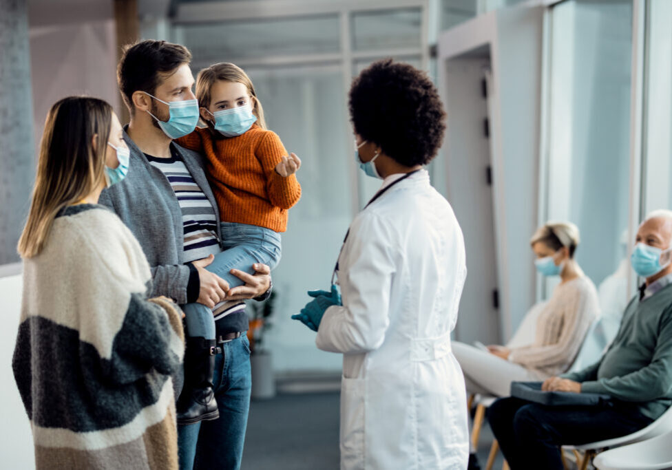 Young parents with small daughter communicating with their family doctor at medical clinic during coronavirus pandemic.