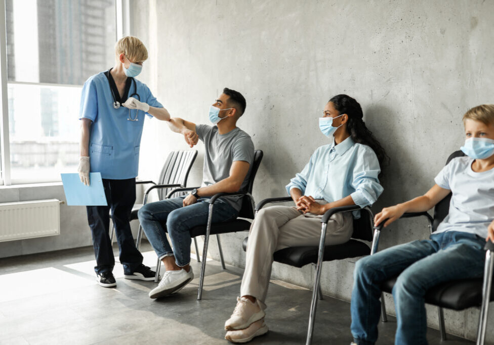 Woman doctor greeting middle-eastern guy patient with elbow bump, multiracial group of people sitting at clinic, waiting for vaccination against COVID-19, wearing protective face masks