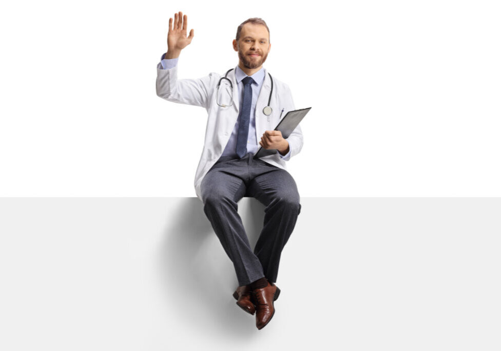 Young male doctor waving and sitting on a blank panel with a clipboard in his hands isolated on white background